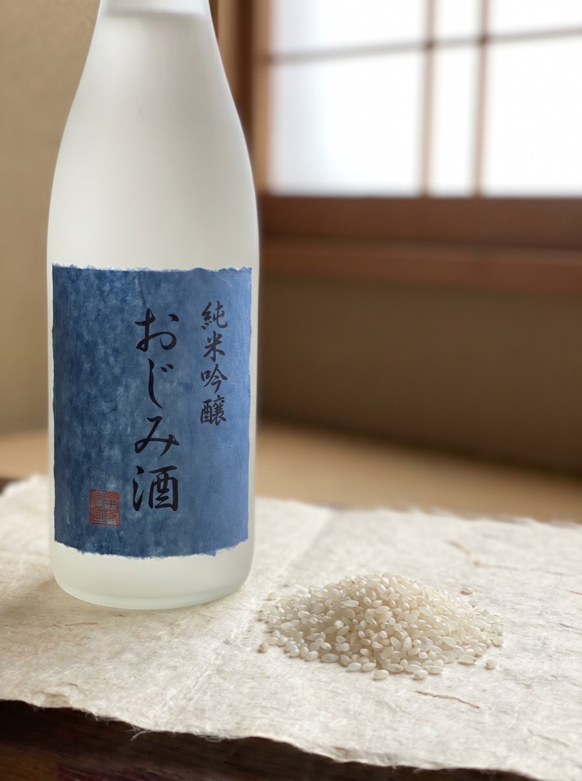 白澤靜物：磨砂白瓶與靛藍標籤置於和紙之上，旁有散落的白米